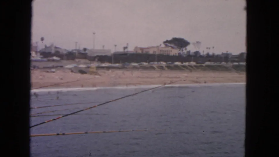 1963 San Francisco frame emphasizing fishing rods over the ocean, with surf, beach activity, buildings, and trees along the shore.