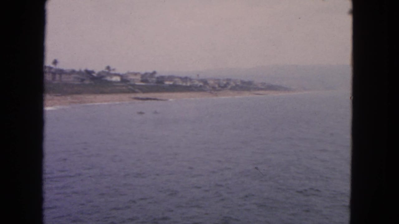 1963 San Francisco-area 8mm frame showing ocean water, a narrow beach, and distant hillside homes under a hazy sky.