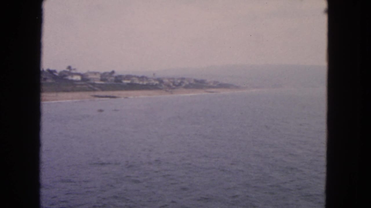 1963 San Francisco-area coastal view with calm ocean, sandy shore, distant houses, and a soft overcast horizon.