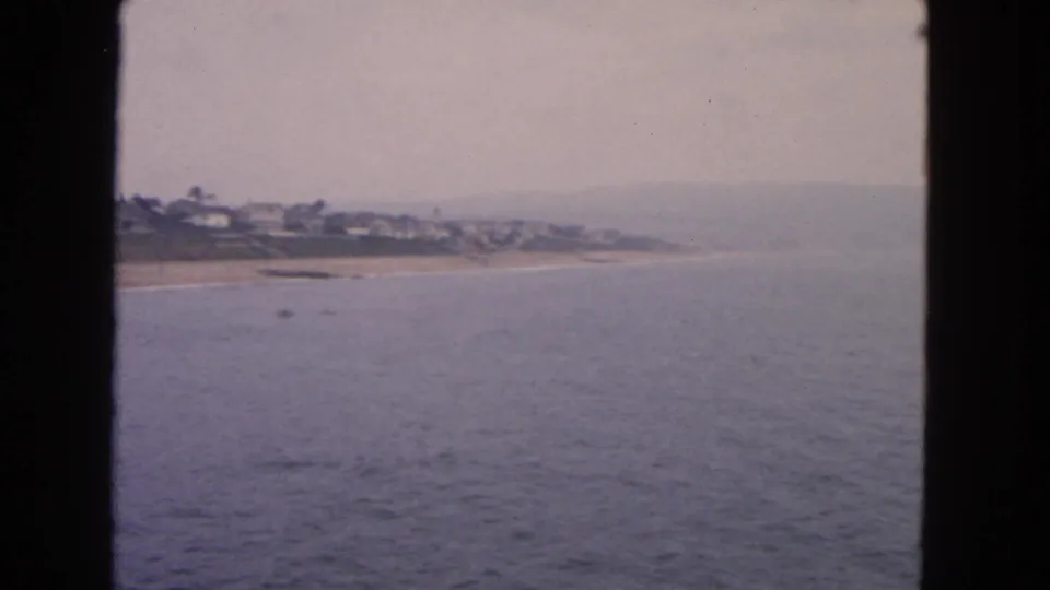 1963 San Francisco-area coastal view with calm ocean, sandy shore, distant houses, and a soft overcast horizon.