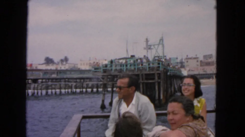 1963 San Francisco home-movie frame of adults on a boat near a pier with water, pilings, and distant buildings.