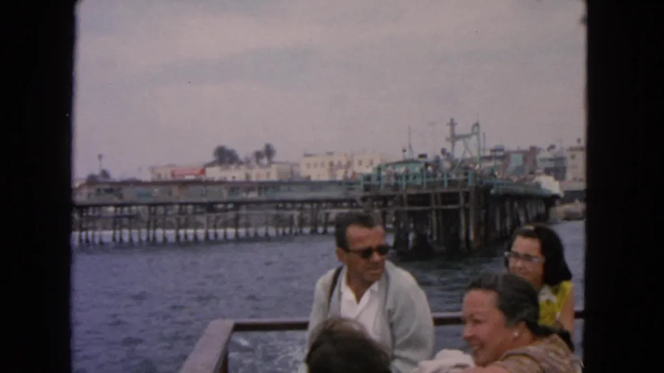 1963 San Francisco boat-ride frame with smiling adults, boat wake, a pier, and distant waterfront buildings.