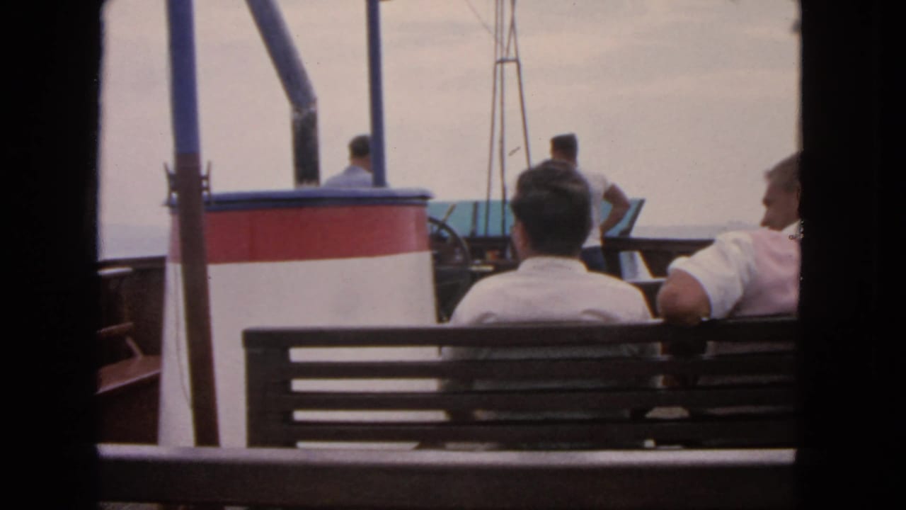 Men seated on a boat bench facing calm water, with a red-and-white cabin structure and blue poles nearby.