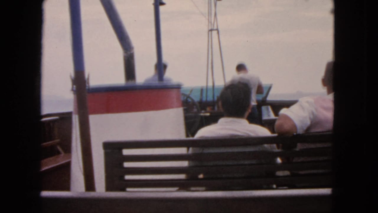 Boat passengers sit with backs to camera while a man stands near the rail and water beyond.
