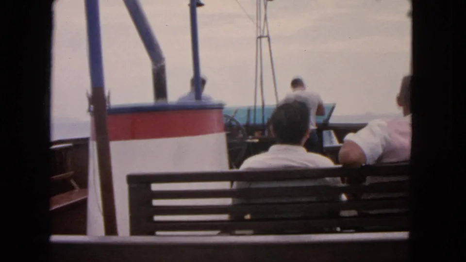 1963 San Francisco boat-trip frame with seated men, a standing man near the rail, and muted overcast water in the background.
