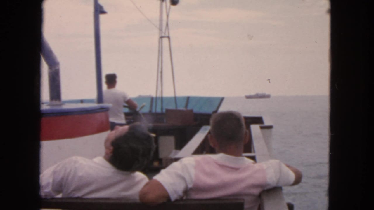 Two seated men and a standing man are seen on a boat with ships in the distance.