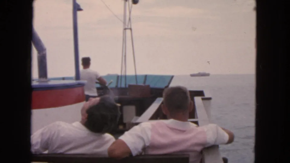 1963 San Francisco frame showing adult men aboard a small boat, with distant ships under an overcast sky.