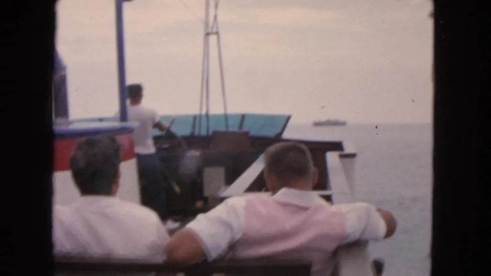 1963 San Francisco 8mm frame of a small boat outing, with men in the foreground and a distant vessel offshore.