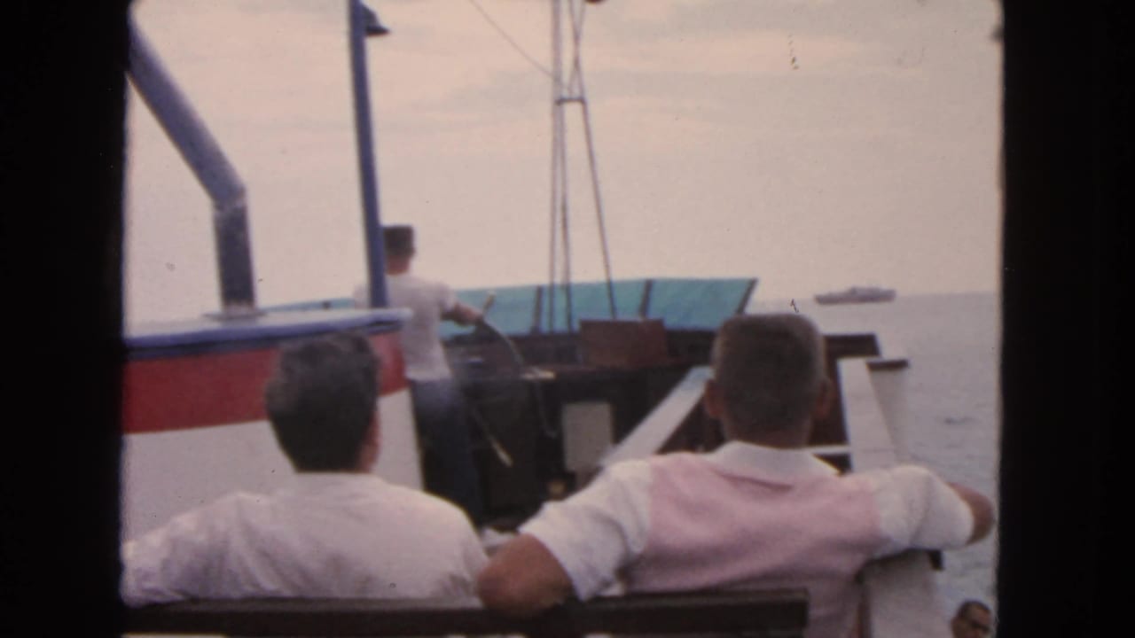 1963 San Francisco home-movie frame showing men seated on a boat deck, open water ahead, and dark film-gate borders at the edges.