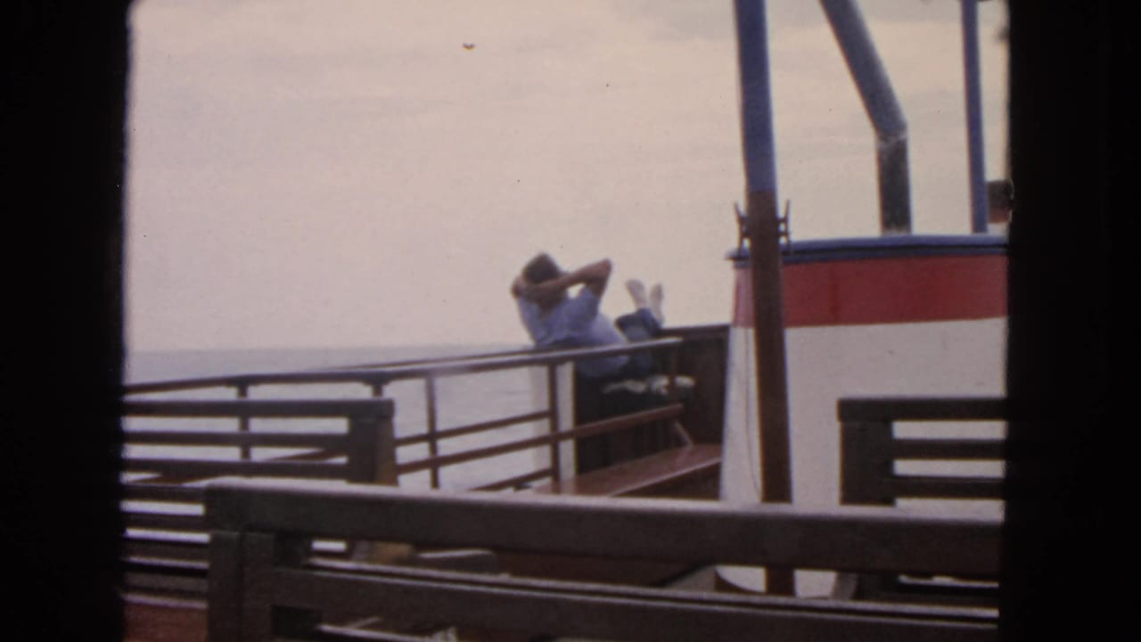 A man reclines on a boat bench with his feet raised beside railings and open water.