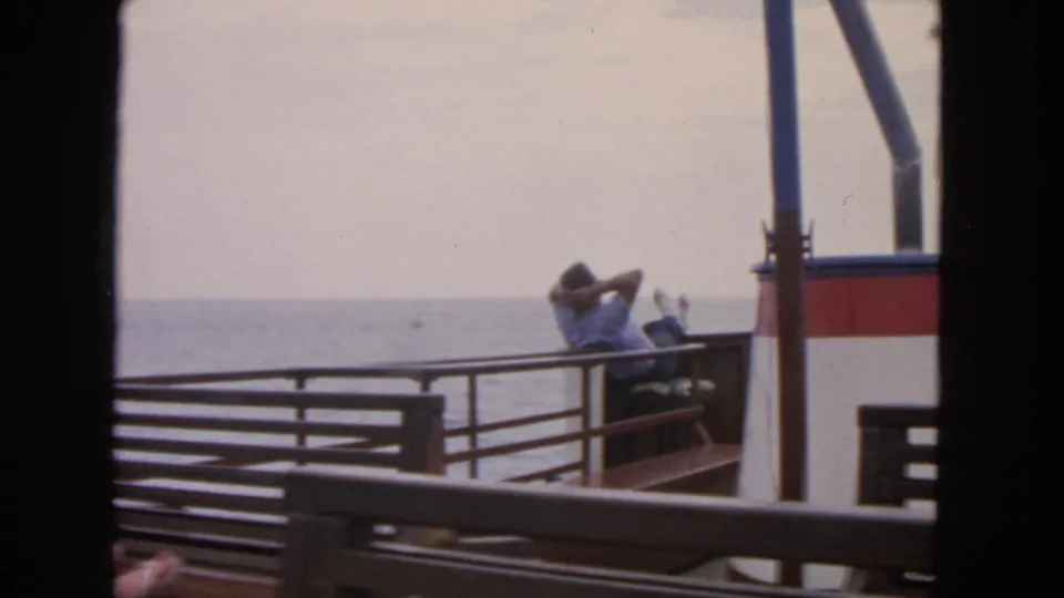 1963 San Francisco home-movie frame showing a reclining passenger on a boat deck framed by railings, benches, and distant water.