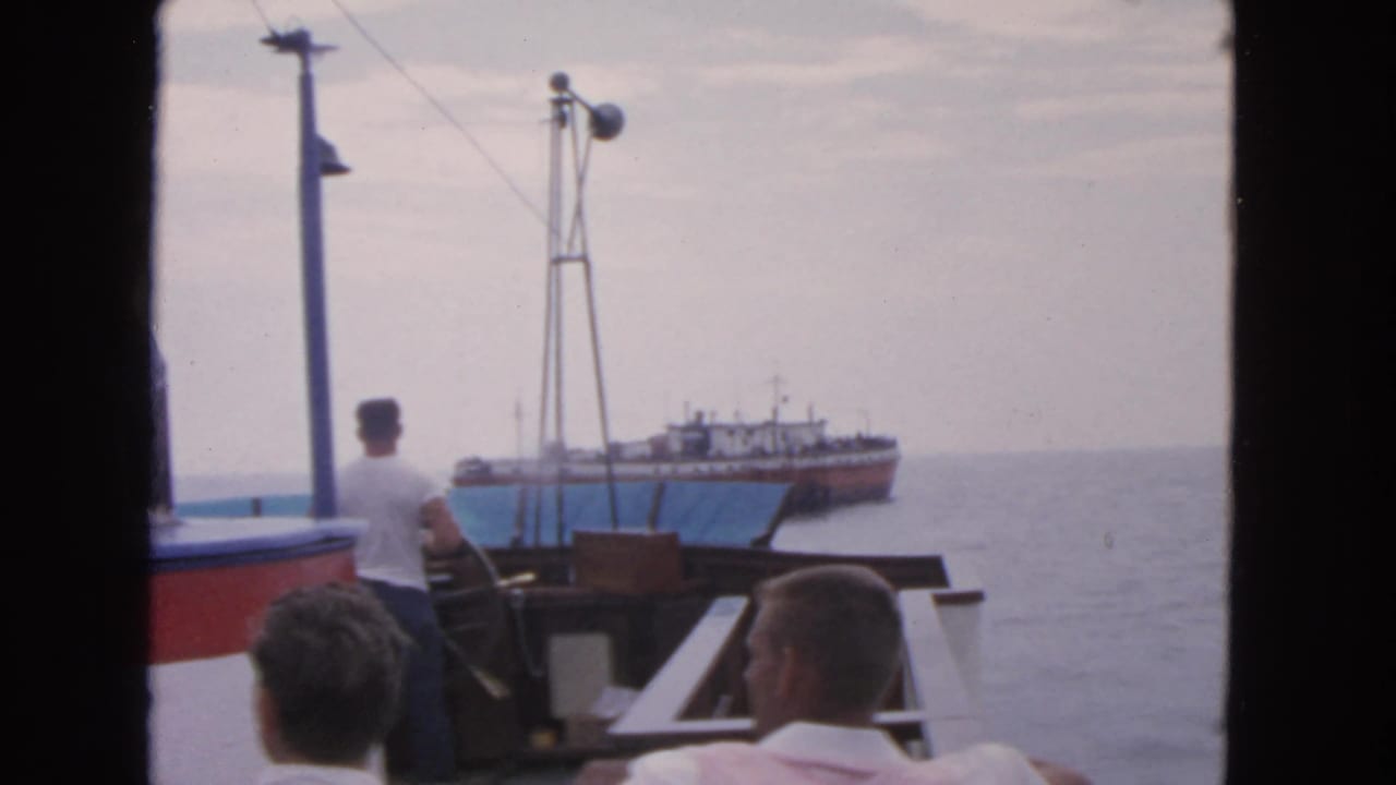 Men sit and stand on a boat deck as a larger ship appears offshore.