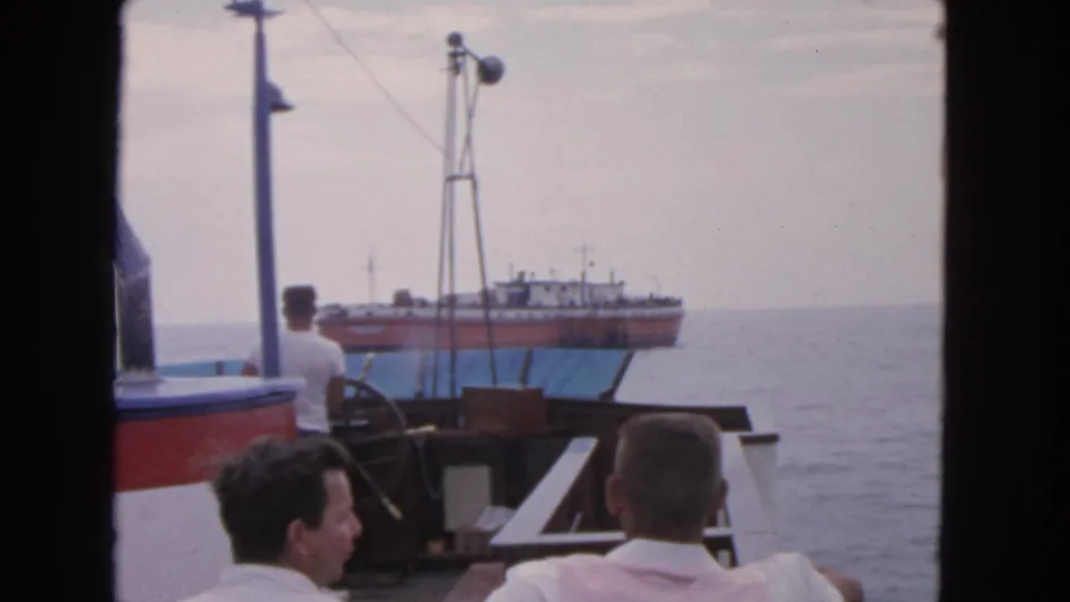Home-movie view from a boat deck with two men in the foreground and a larger ship beyond the railing.