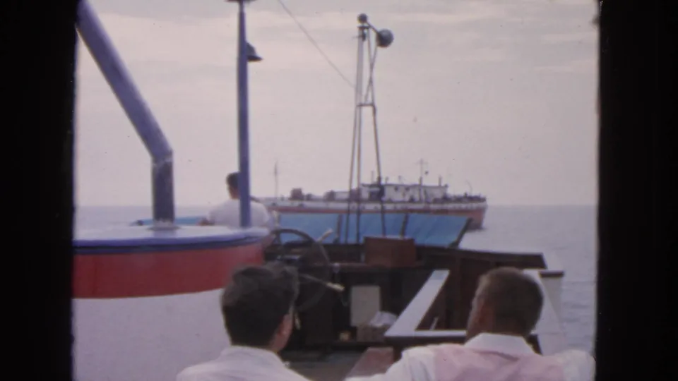 1963 San Francisco boat-trip frame with men on deck, mast hardware, bay water, and a larger ship offshore.