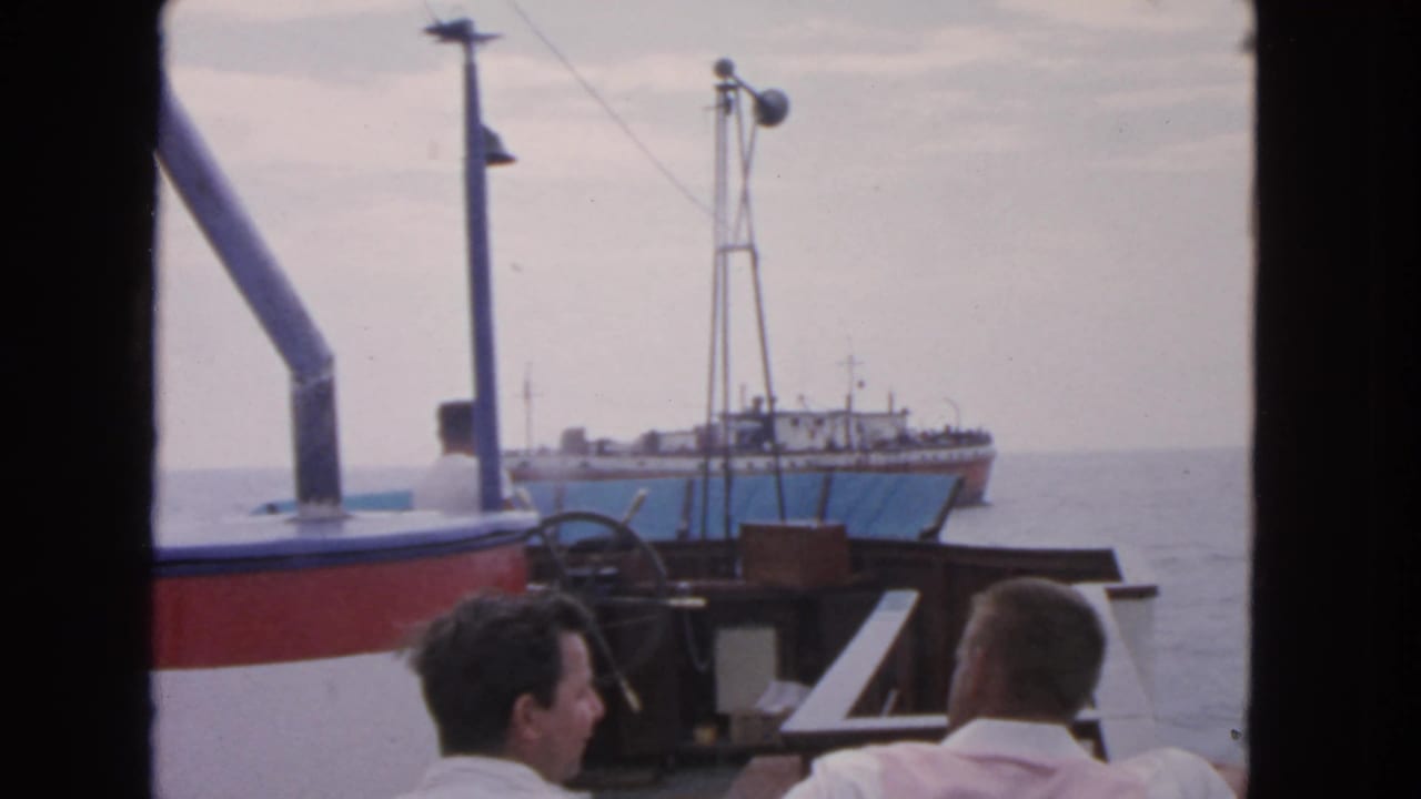 Two men sit on a boat deck as another vessel appears ahead on open water.