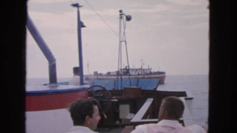 1963 San Francisco home-movie frame showing two men on a boat deck approaching a blue-and-white vessel on the water.