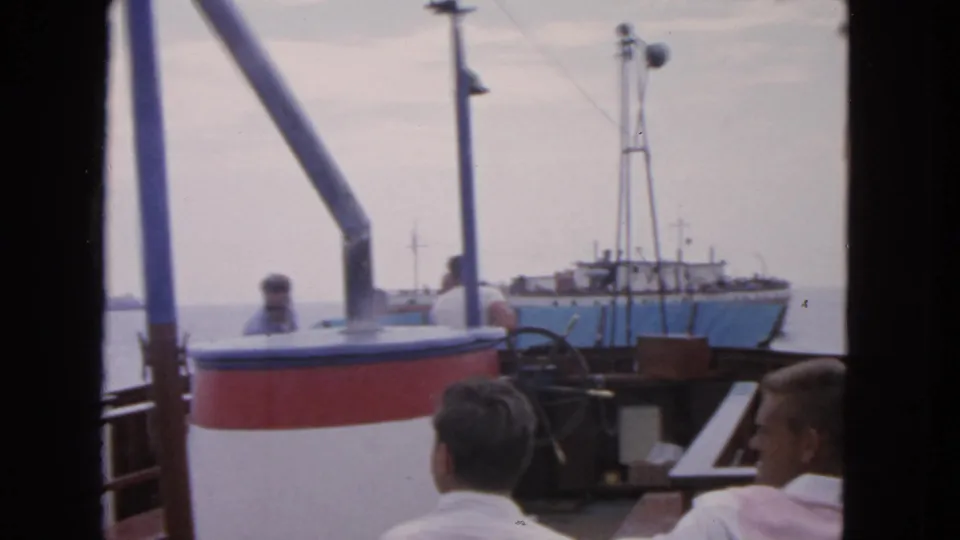 1963 San Francisco 8mm frame showing a boat deck, seated men, and a nearby blue-and-white vessel on open water.