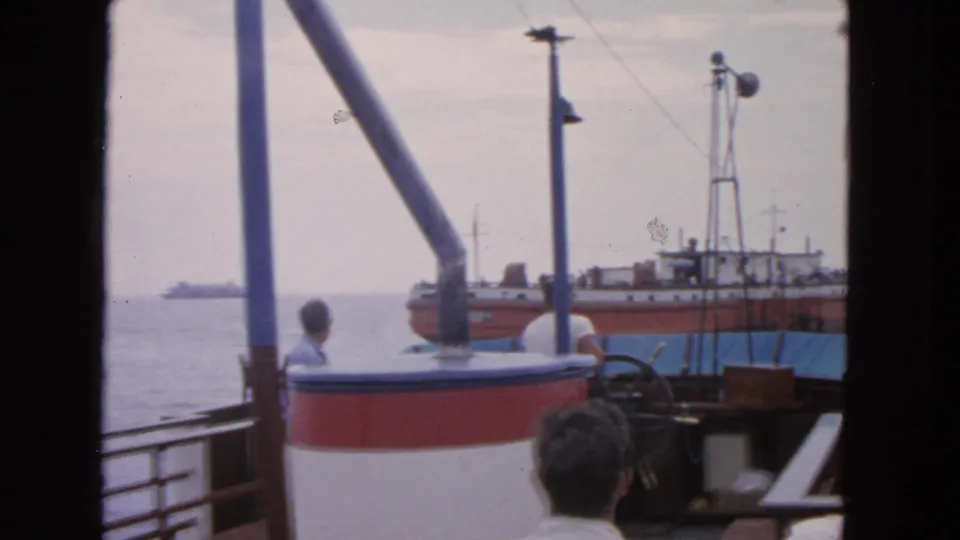 1963 San Francisco home-movie still of a seated passenger on a boat deck with a larger vessel close behind.