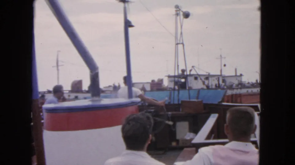 1963 San Francisco frame showing men on a boat deck, one near the wheel, with a large fishing vessel across the water.