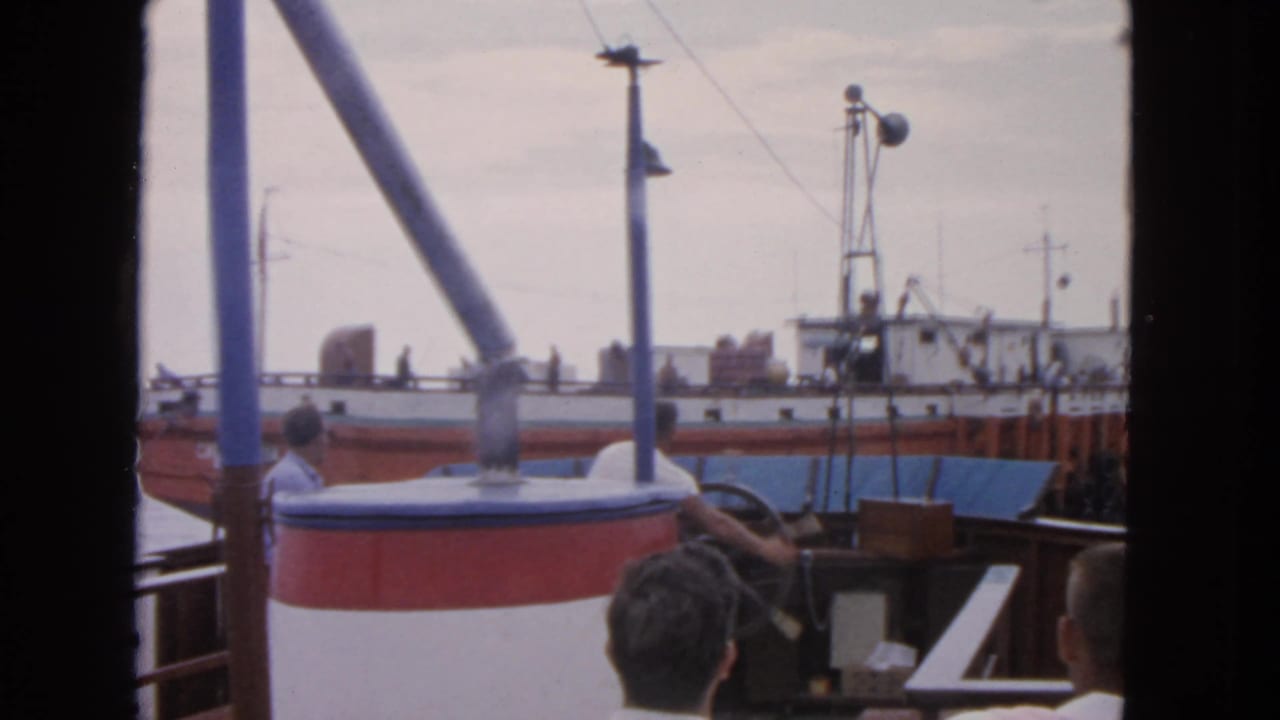 Passengers on a boat deck look toward a fishing vessel across the harbor.