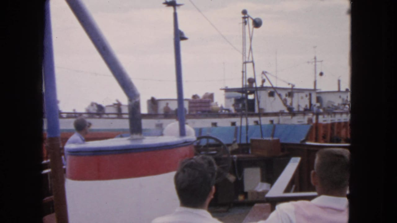 Two passengers sit in the foreground as a fishing vessel fills the harbor view.