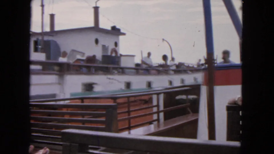 8mm frame of a white passenger boat passing close by, with adults on deck and another vessel edge in the foreground.