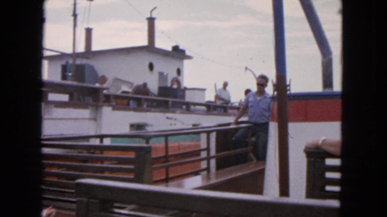 Passengers stand on the deck of a white vessel seen past railings from another boat.
