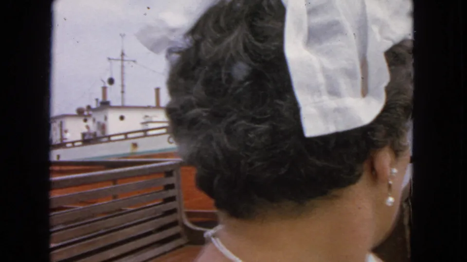 1963 San Francisco ferry frame showing a woman seen from behind beside wooden benches as another vessel passes beyond the railing.