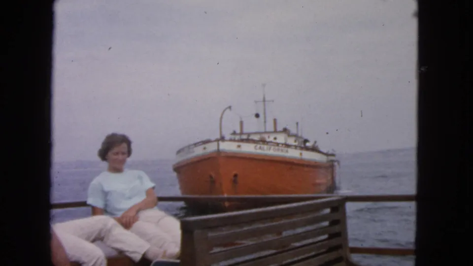 1963 San Francisco Bay home-movie still of a woman seated on deck with the ship CALIFORNIA and hazy shoreline behind her.