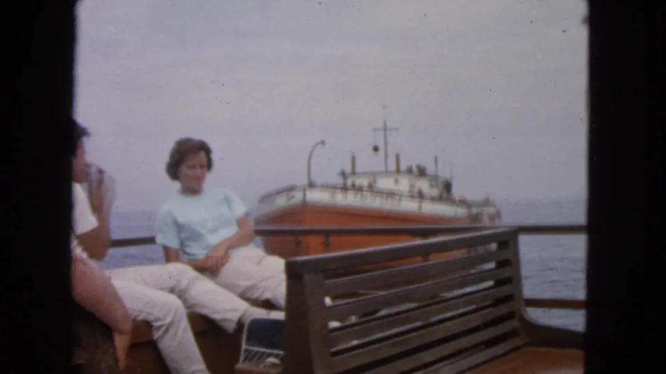 1963 San Francisco-area home-movie frame showing adults seated near a boat railing as an orange and white ship passes offshore.