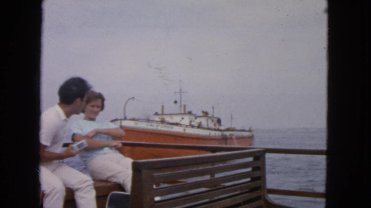 A man and woman sit beside the railing on a boat deck in 1963 San Francisco-area footage, with a named orange and white vessel behind them.