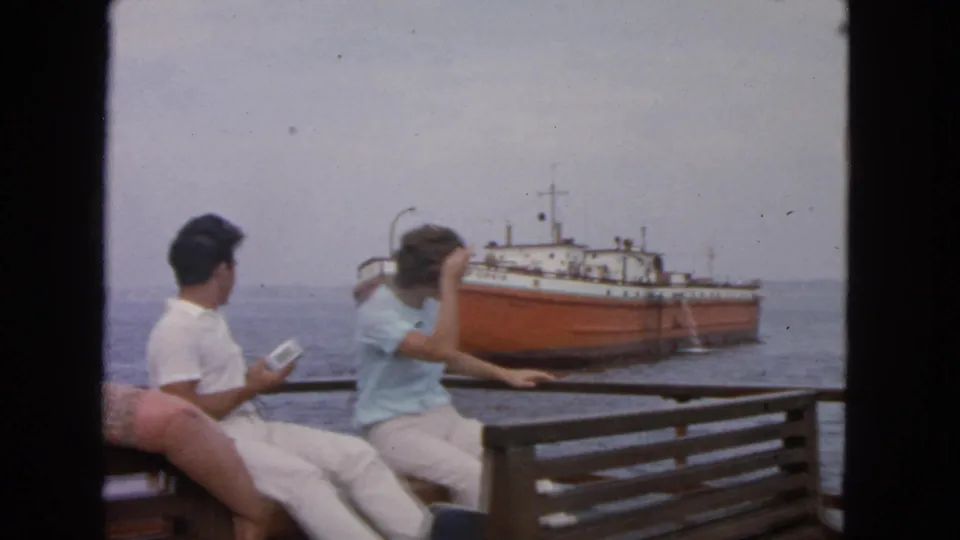 The woman gestures near her hair while seated with a man on a boat deck, facing an orange and white ship in supported 1963 San Francisco footage.