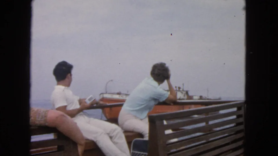 On a boat deck in 1963 San Francisco-area footage, a woman leans forward toward the rail as a man watches the orange and white vessel nearby.