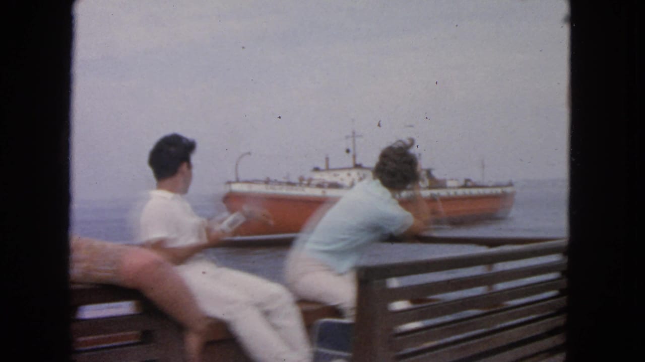 1963 San Francisco Bay home-movie frame with passengers seated by a wooden bench as a red-hulled ship passes in the distance.