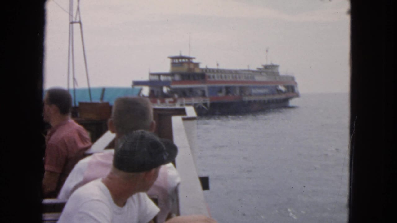 Passengers on a boat look toward a multi-deck ferry on open water.