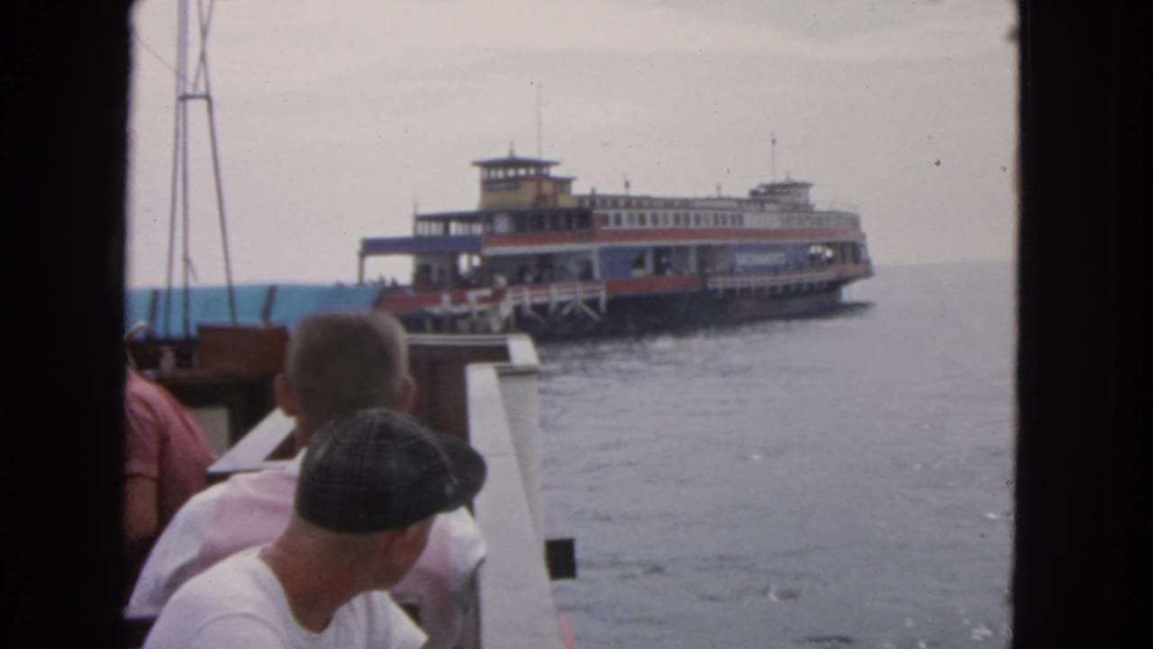 1963 San Francisco bay view with a red, white, blue, and yellow ferry seen past passengers and boat railings.