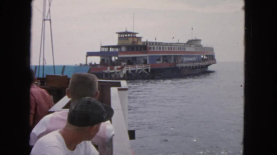 1963 San Francisco home-movie frame with seated passengers watching a multi-level ferry under a pale sky.