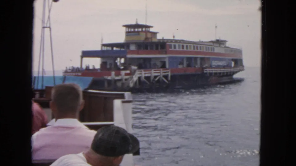 The ferry appears closer to the dock, with foreground passengers facing the water from another boat in San Francisco.