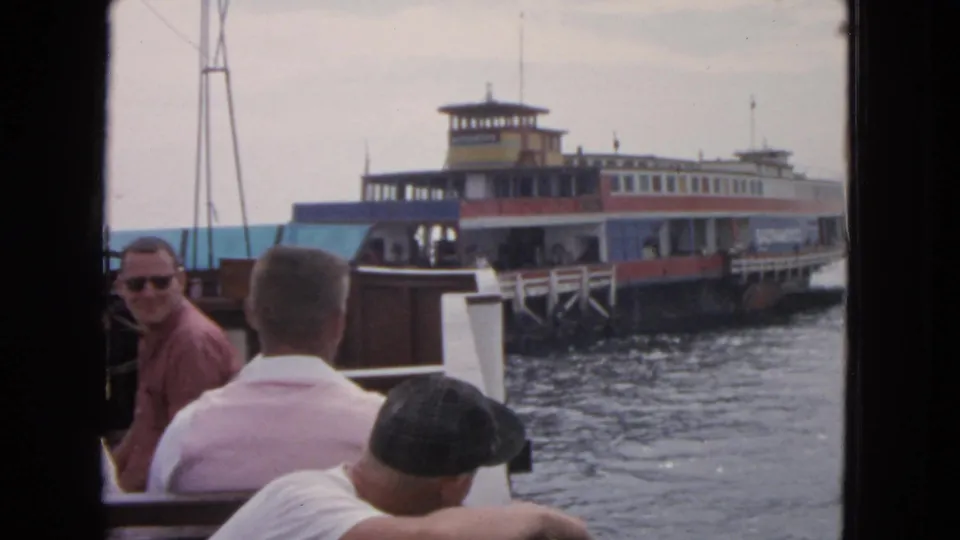 A boat passenger in sunglasses looks back toward the camera while the 1963 San Francisco ferry nears the dock behind him.