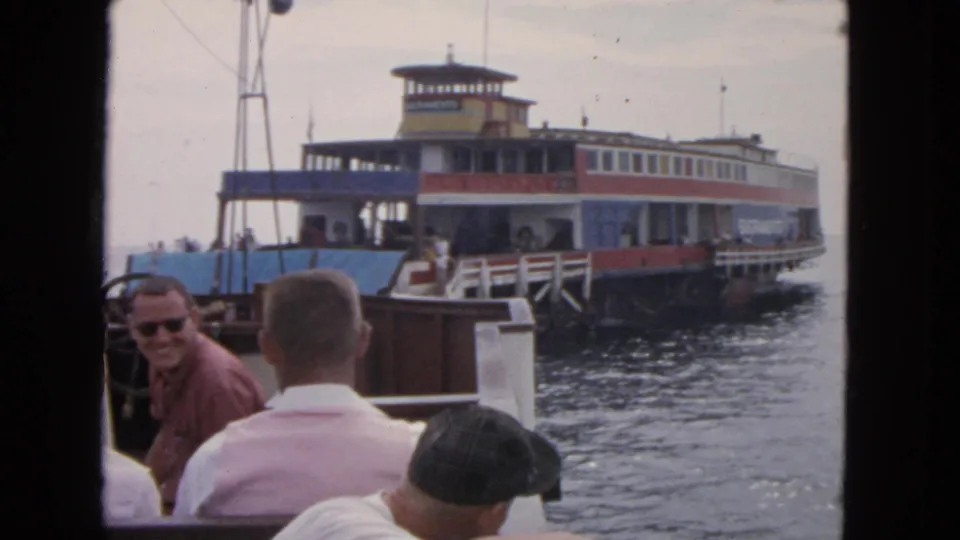 The ferry is close to the dock, with seated boat passengers in the foreground and water between the two vessels.
