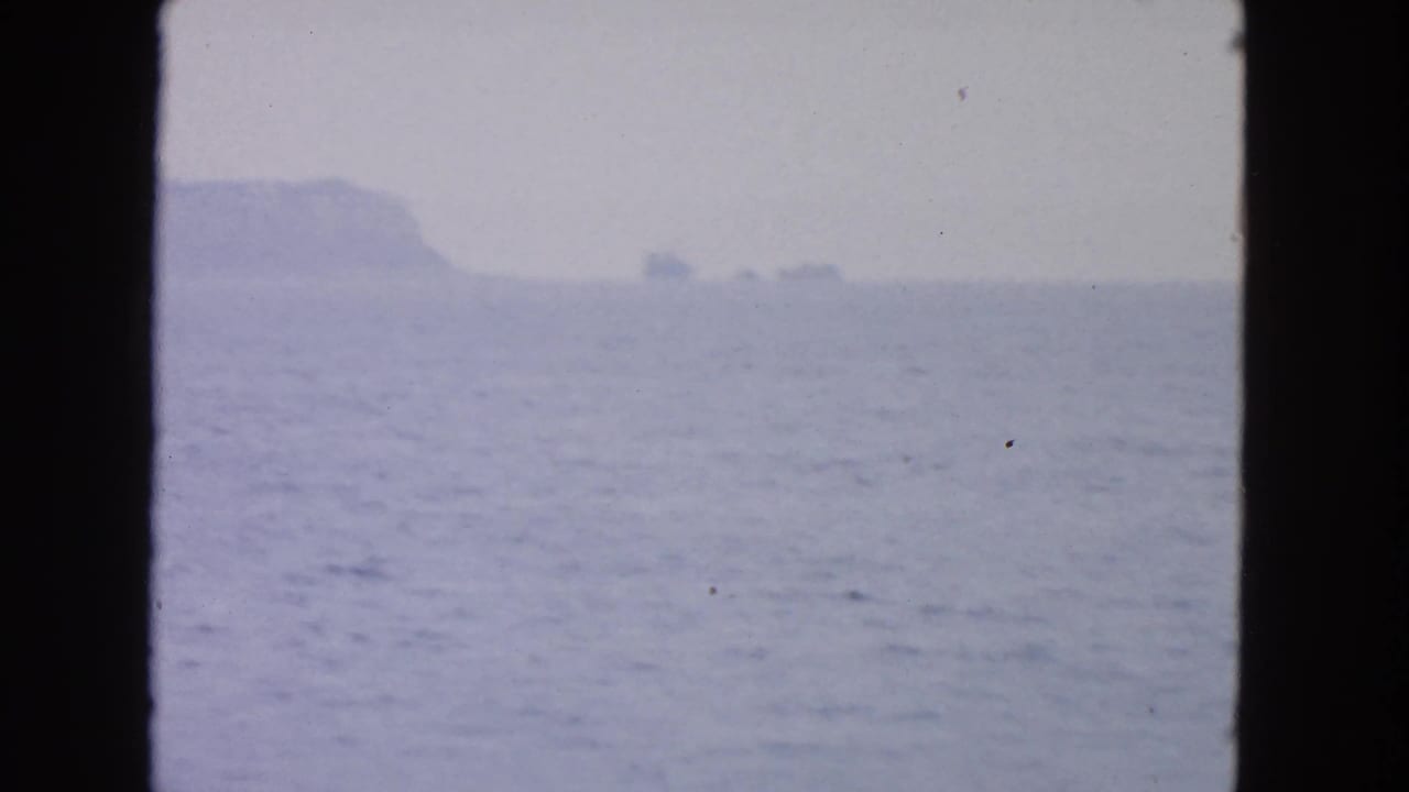 1963 San Francisco Bay-area home-movie frame showing hazy water and a distant island or cliff through dark boat-window framing.
