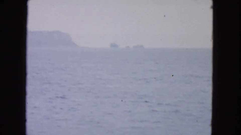 1963 San Francisco Bay-area home-movie frame showing hazy water and a distant island or cliff through dark boat-window framing.