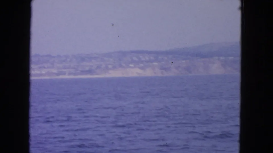 1963 8mm San Francisco-area coastal view from a moving vessel, with blue water, distant hills, and a hazy shoreline framed by dark edges.