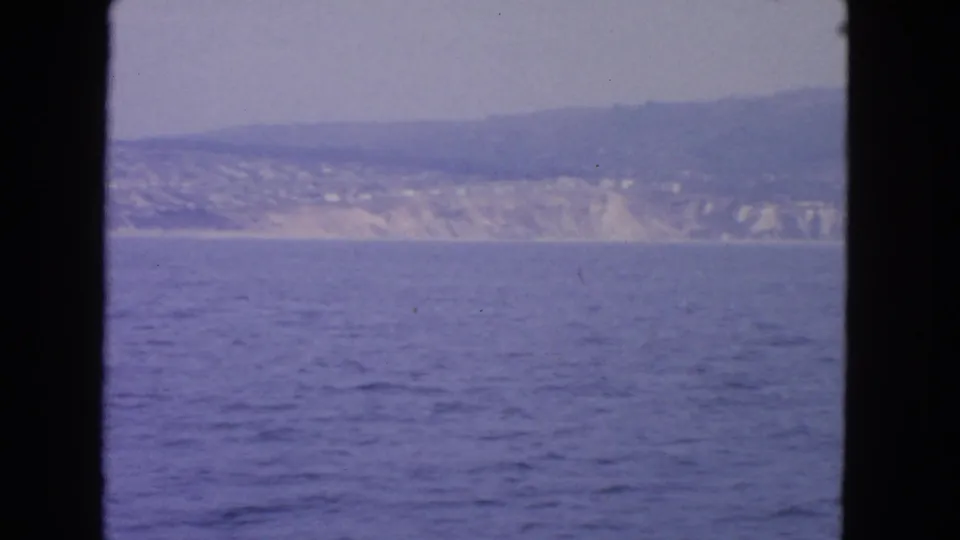 1963 San Francisco-area home-movie frame of blue coastal water, pale shoreline cliffs, and a hazy hillside seen from a moving vessel.
