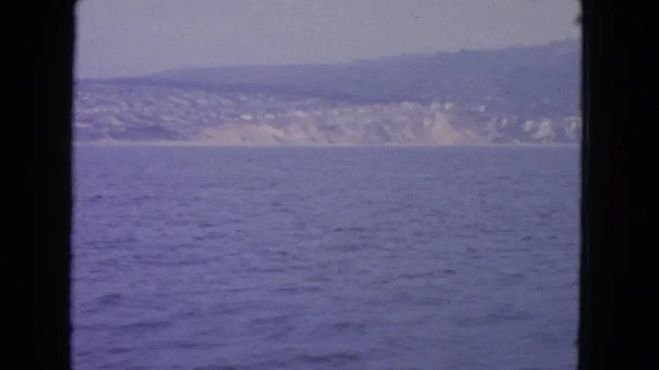 1963 8mm view from offshore toward a hazy California coastline with blue water, low hills, and distant buildings.
