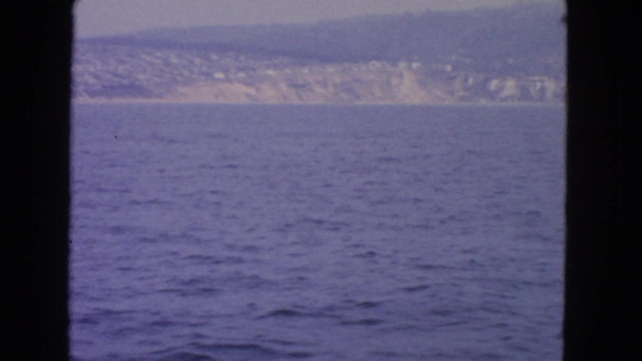 Boat-view 8mm frame showing open water and a pale, hilly California shoreline under a muted sky.