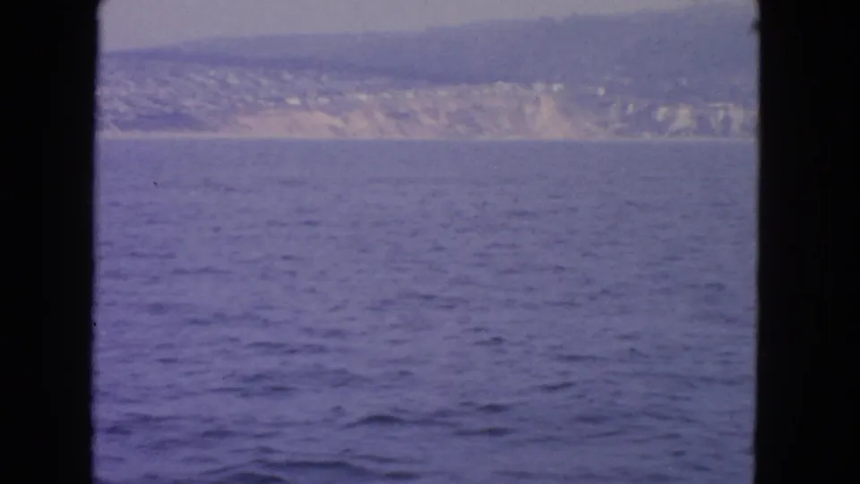 Boat-view 8mm frame showing open water and a pale, hilly California shoreline under a muted sky.