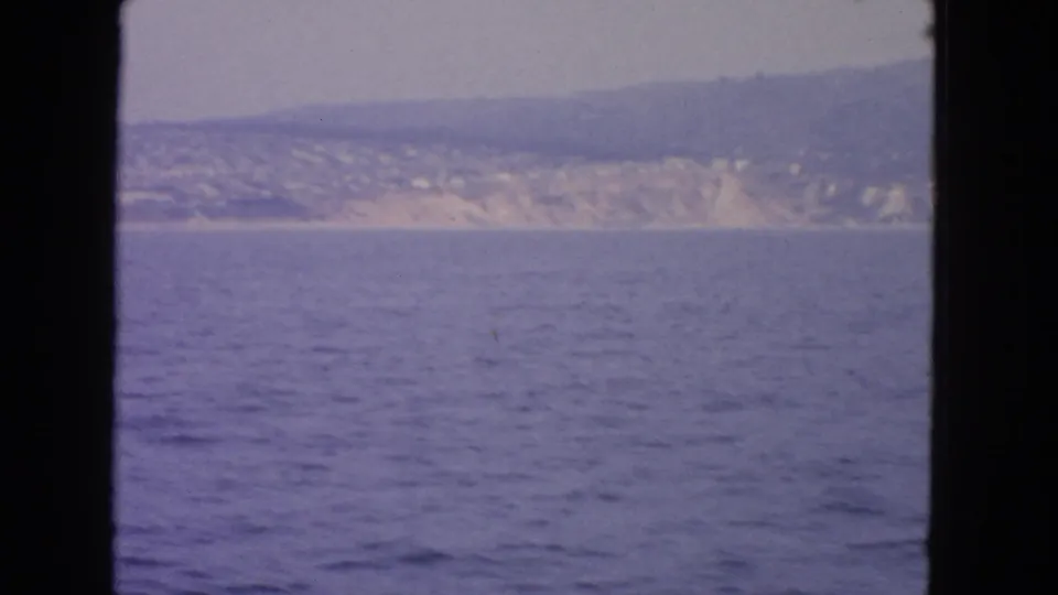 1963 home-movie frame with blue-purple water in front of a distant coast and softened hillside detail.