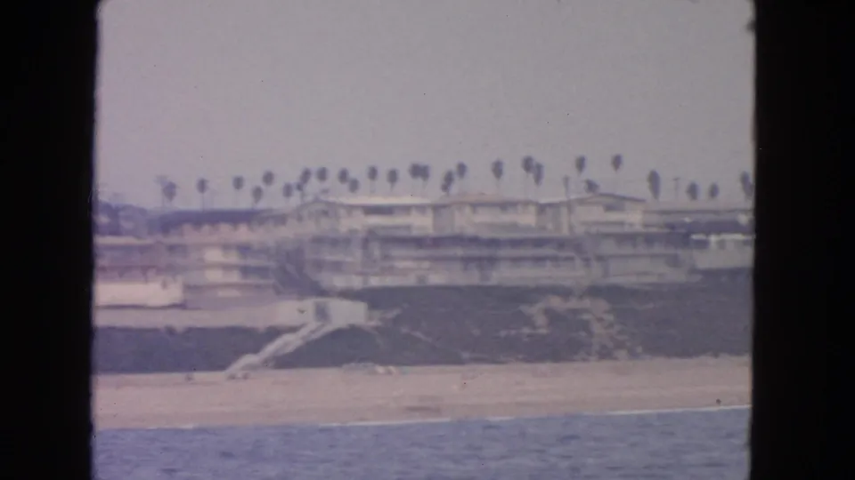 1963 coastal frame showing a beachside structure, rows of palms, sand, and water from an offshore viewpoint.