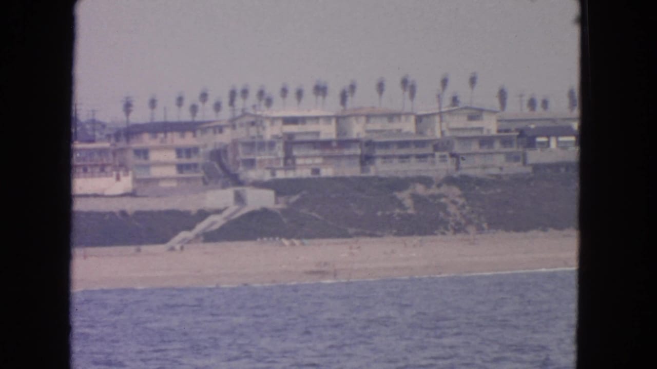 Ocean view toward a sandy beach, grassy bluff, palm trees, and seaside apartment buildings.